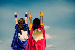 two girls wearing capes with their arms stretched high, and a sky background